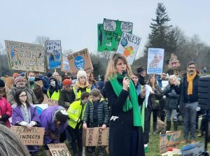 Jo Robb in a black coat and green scarf, standing in front of a crowd holding placards with slogans such as "Cut the Crap", "Save Our Rivers, Stop Poollution"